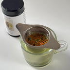 Clear glass cup with tea infuser, filled with Genmaicha tea leaves, on a white background.