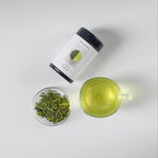 Green tea leaves in a glass bowl, brewed tea in a cup, and a tea tin product on a light gray background