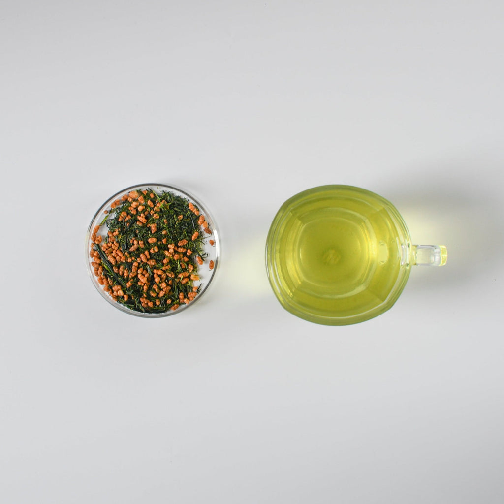 Clear mug with brewed green tea and small petri dish with genmaicha loose leaf green tea on a white background