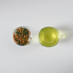 Clear mug with brewed green tea and small petri dish with genmaicha loose leaf green tea on a white background