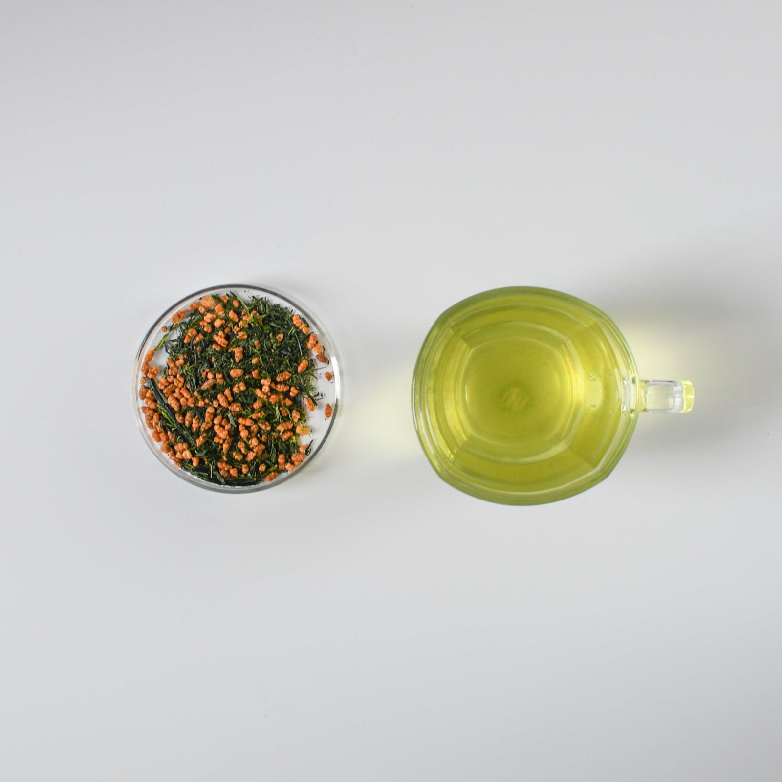 Clear mug with brewed green tea and small petri dish with genmaicha loose leaf green tea on a white background